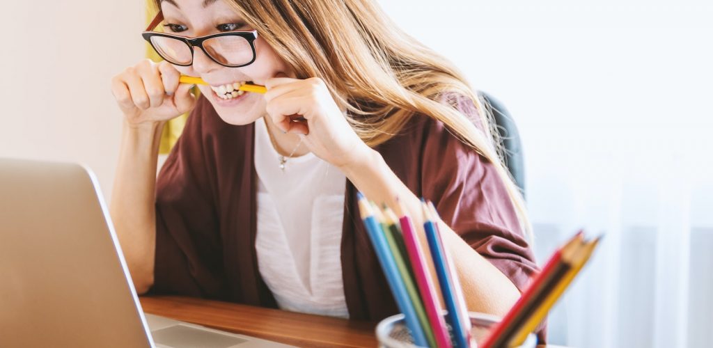 woman biting pencil while sitting on chair in front of computer during daytime