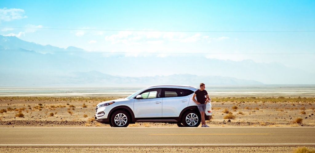 man standing beside white SUV near concrete road under blue sky at daytime