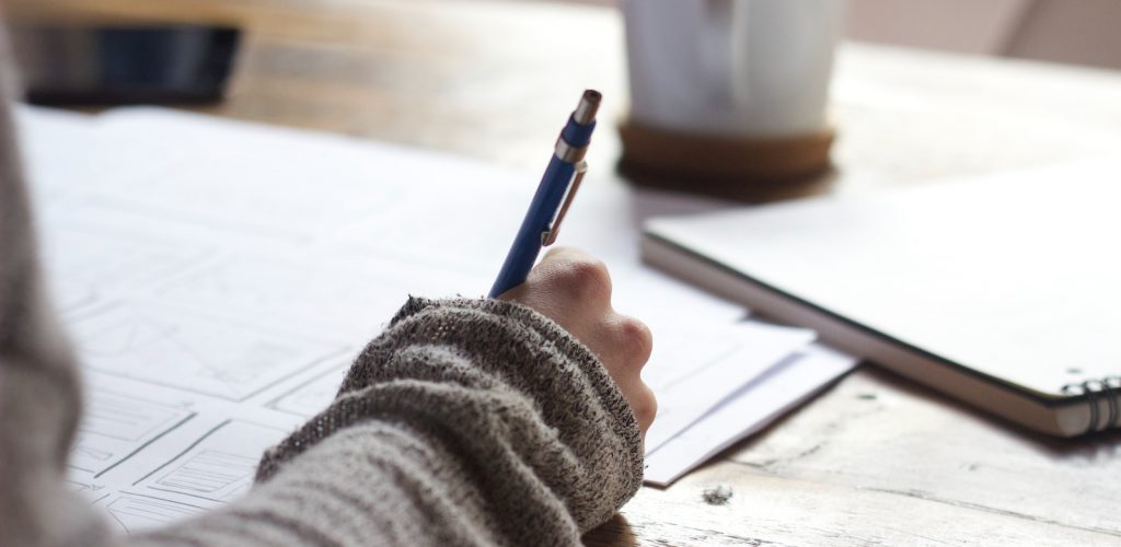 person writing on brown wooden table near white ceramic mug